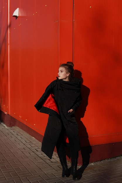 fashionable teenage girl wearing black warm clothing standing against red wall during sunny day