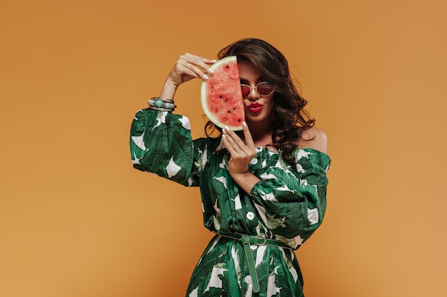 Fashionable wavy dark haired girl with red lipstick and white manicure posing in green modern dress on isolated background and covers face with piece of watermelon