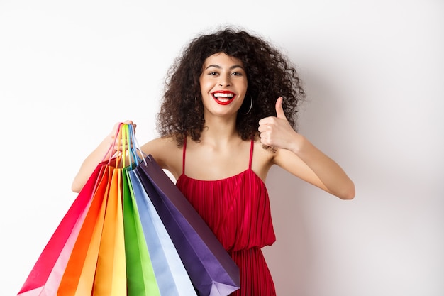 Fashionable woman in red dress going shopping, holding bags and showing thumbs up, recommend store, standing on white background.