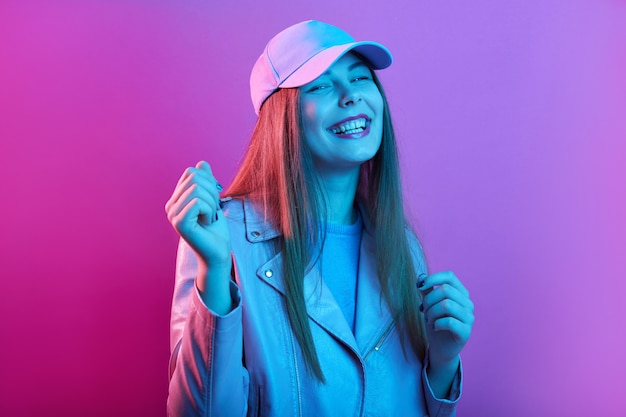 Fashionable woman stands smiling against pink neon wall, having fun in stylish outfit