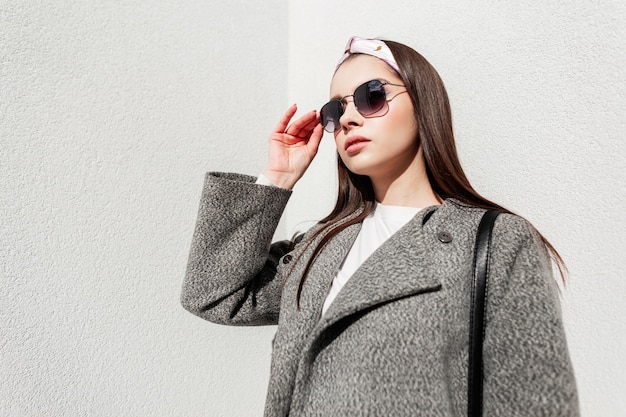 Fashionable woman in stylish spring clothes straightens sunglasses near wall on sunny day. Cute girl in trendy outfit with glasses in bandana poses on bright sunny day on street in city. Youth style.