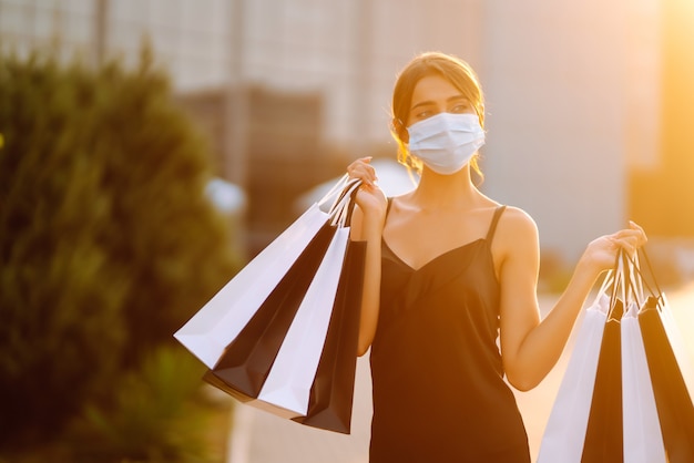 Fashionable woman wearing protective medical mask with shopping bags at sunset.