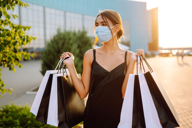 Fashionable woman wearing protective medical mask with shopping bags at sunset.