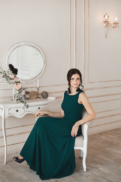 Fashionable woman with stylish hairstyle in evening dress and in trendy shoes sits at the dressing table