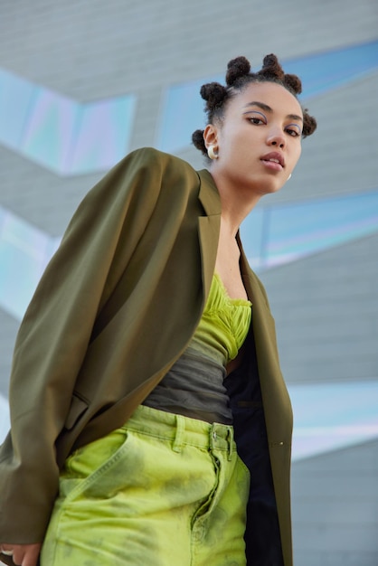 Fashionable woman with trendy hairstyle dressed in green fashionable clothes looks confidently at camera wears blue eyeliner poses against blurred background View from top People and style