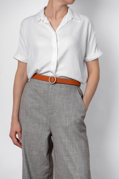 Fashionable women's casual spring outfit woman posing in a white blouse and gray trousers with belt closeup faceless hands in pockets business style