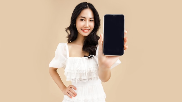 ファッショナブルな若いアジア女性のスマート フォンを使用して孤立したベージュ色の背景に立っている幸せを感じて携帯電話でショッピング オンライン決済携帯電話で空白の画面の携帯電話を示す女性