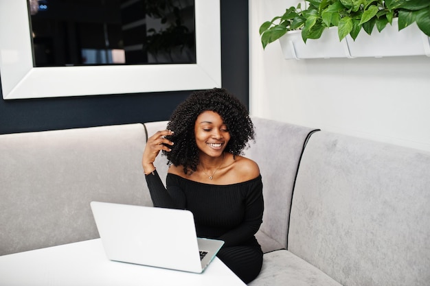 아프리카 헤어스타일을 한 세련된 젊고 아름다운 아프리카계 미국인 여성 사업가는 우아한 검은색 옷을 입고 노트북에서 일하고 있습니다.