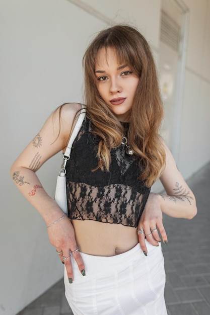 Fashionable young beauty woman with cute face in stylish sexy lace top and black bra with skirt and handbag walks on the street near a gray wall