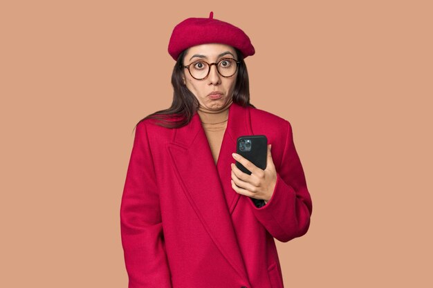 Photo fashionable young caucasian woman with phone in red coat and beret on studio background