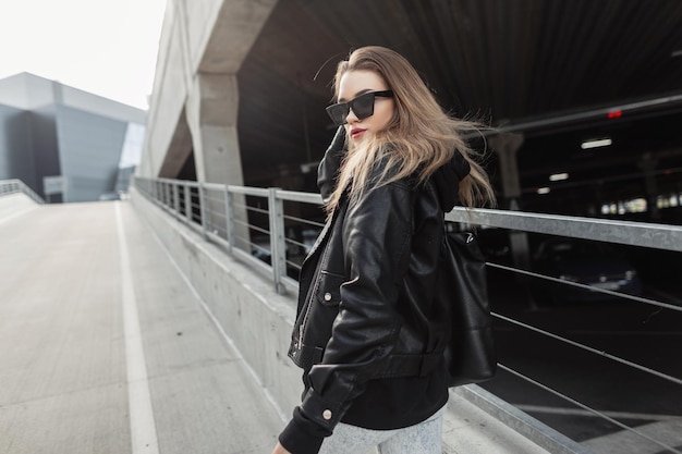 Fashionable young girl with cool fashion sunglasses in trendy black leather clothes with leather rock jacket and bag walks in the city Urban female spring style outfit