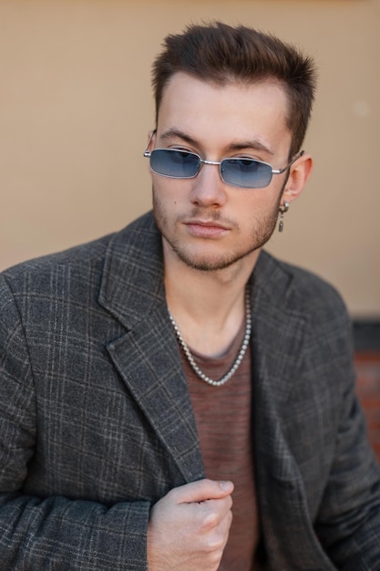 Fashionable young guy hipster with blue sunglasses in trendy coat sits in the city