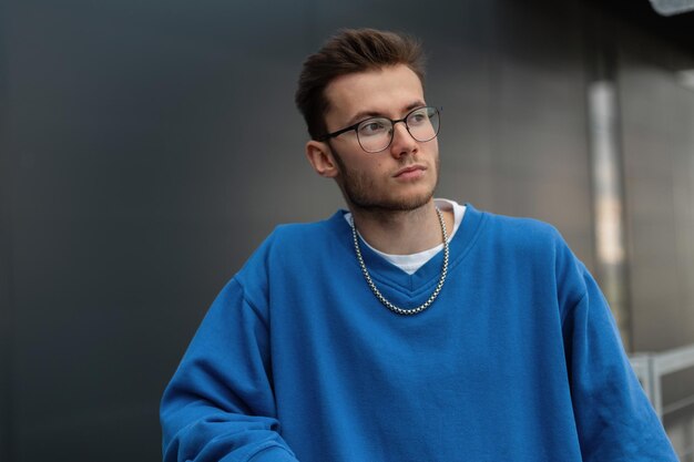 Fashionable young handsome hipster guy with glasses in a stylish blue sweatshirt in the city
