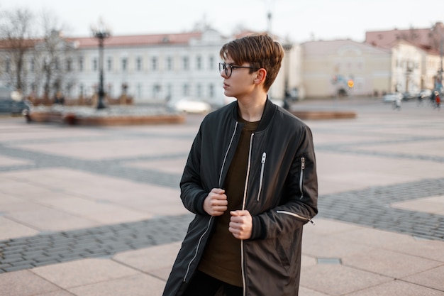 Fashionable young hipster man in stylish glasses with a stylish hairstyle in a fashionable long black jacket in a T-shirt posing in the city outside on a warm autumn day. Cool guy for a walk.