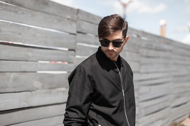 Fashionable young man in a black jacket in dark sunglasses enjoys a walk on the street on a sunny summer day. American stylish guy relaxes near a vintage wooden fence outdoors.