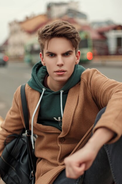 Fashionable young man with a haircut in a vintage coat with a hoodie and a black bag on the street