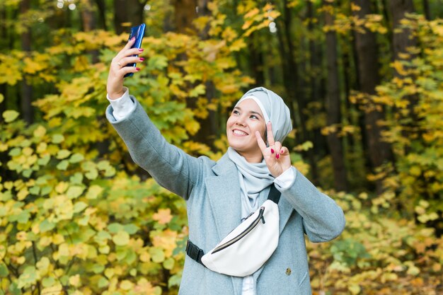 秋の公園で屋外のスマートフォンでselfieを取るヒジャーブのファッショナブルな若いイスラム教徒のアジアの女の子