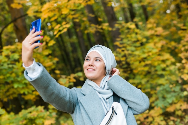 秋の公園で屋外のスマートフォンでselfieを取るヒジャーブのファッショナブルな若いイスラム教徒のアジアの女の子