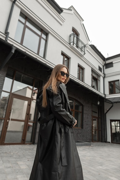 Fashionable young pretty woman with vintage sunglasses in stylish black long leather coat walks near a building on the street