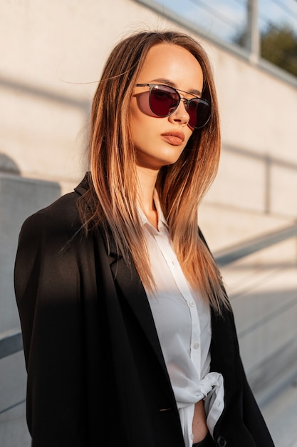 Fashionable young woman model in trendy sunglasses in fashion casual black jacket in beauty stylish white shirt near vintage wall on summer sunny day. Cool modern girl in youth clothes on sun in city.