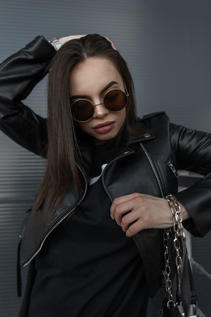 Fashionable young woman model with cool vintage sunglasses and stylish leather jacket and black dress with fashion bag looks dawn near metal wall on the street