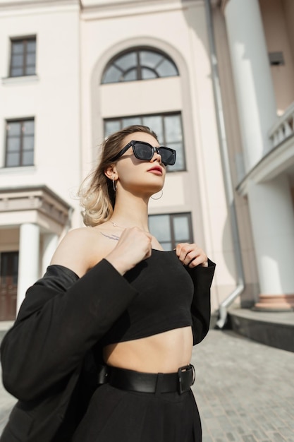 Modello di giovane donna alla moda con occhiali da sole alla moda in abiti eleganti da lavoro neri cammina per strada vicino a un edificio vintage