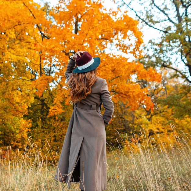 세련된 가을 옷을 입은 세련된 젊은 여성이 공원에서 가을 풍경을 즐깁니다. 세련된 모자에 트렌디 한 긴 코트에 우아한 소녀가 숲에 서 있습니다. 뒤에서 봅니다.