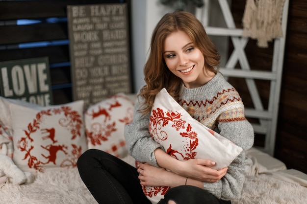 Fashionable young woman in a stylish knitted sweater with a pillow sits on the bed