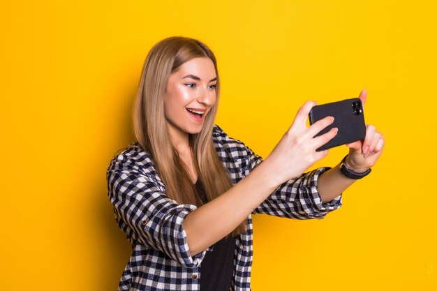 La giovane donna alla moda prende selfie sul telefono che posa sulla parete gialla,