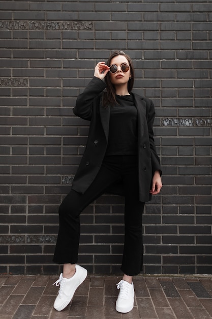 Fashionable young woman in vintage black blazer in fashion t-shirt in stylish jeans pants stands near black brick wall in city. Close-up of female body. Spring collection of women's casual clothing.