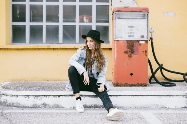 Fashionable young woman wearing hat sitting beside an old petrol pump