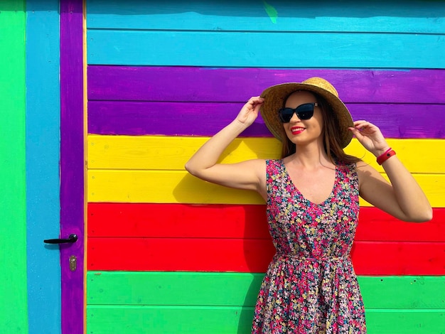 Foto giovane donna alla moda che indossa un cappello di paglia e occhiali da sole in piedi contro un muro luminoso e colorato