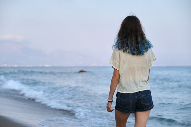 Giovane donna alla moda con capelli blu lunghi tinti che cammina lungo la spiaggia. bellezza, moda, concetto di natura. vista posteriore, tramonto sul mare, copia dello spazio