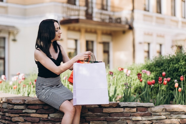 Fashionably dressed woman with colored shopping bags sitting near flowers on the streets, shopping concept