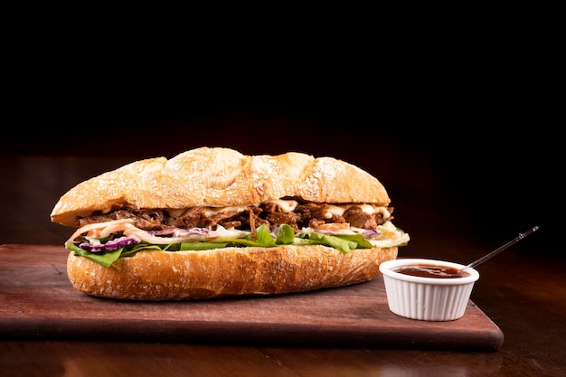 Fast food beef brisket sandwich with arugula and coleslaw salad on baguette bread on wooden board with barbecue sauce dark background angle