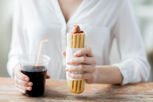 fast food, people and unhealthy eating concept - close up of woman hands with hot dog and coca cola drink sitting at table