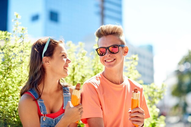 fast food, summer and people concept - happy teenage couple eating hot dogs sitting on city street bench