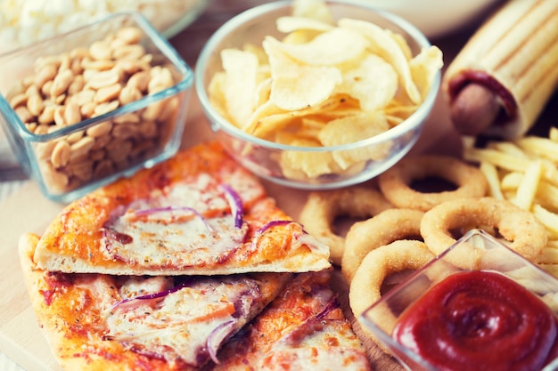 fast food and unhealthy eating concept - close up of pizza, deep-fried squid rings, potato chips, peanuts and ketchup on wooden table top view