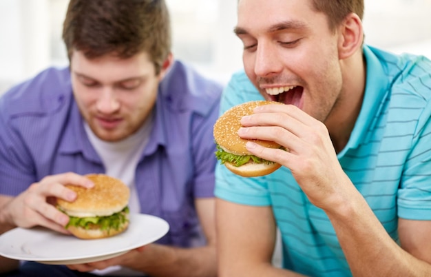 Photo fast food, unhealthy eating, people and junk-food - close up of happy friends eating hamburgers at home