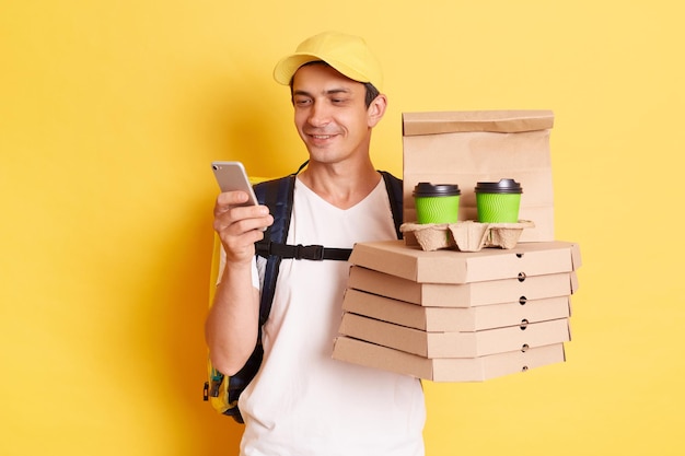 Fast service and best food delivery Smiling Caucasian delivery man wearing cap and white t shirt holding pizza boxes and takeaway coffee and using mobile phone isolated on yellow background