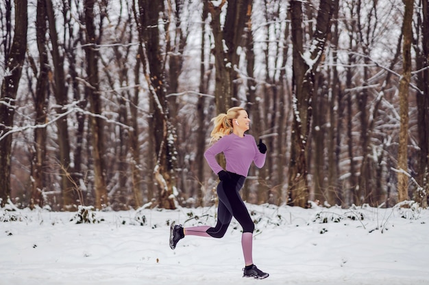 Fast sportswoman in warm sportswear running in woods in snow in the nature at winter. Cardio exercises, winter fitness, cold weather