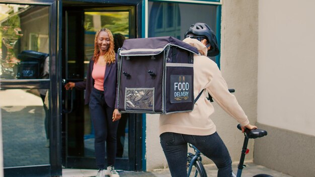 Fastfood-bezorger die maaltijdbestelling in pakket geeft aan afro-amerikaanse vrouw, die papieren zakvoedsel bezorgt bij de voordeur. Vrouwelijke koerier met afhaal fastfood lunch, expresdienst.