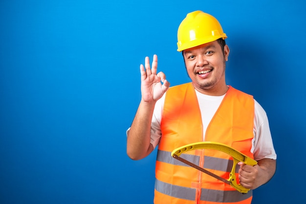 Fat asian construction worker wearing orange safety vest and helmet showing okay sign while holding hacksaw