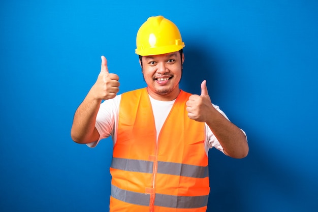 Fat asian construction worker wearing orange safety vest and helmet showing thumb up sign smiling