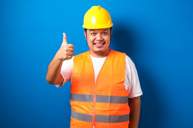 Fat asian construction worker wearing orange safety vest and helmet showing thumb up sign smiling