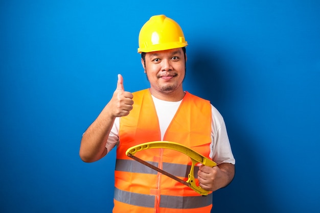 Fat asian construction worker wearing orange safety vest and helmet showing thumb up sign while holding hacksaw