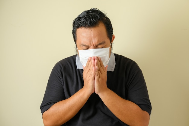 Толстый азиатский парень в маске кашляет, закрыв рот руками, плохо себя чувствует. симптомы болезни коронавируса