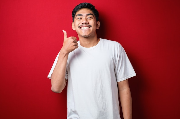 Fat Asian guy wearing a white T-shirt smiles at the camera while showing his thumbs up. Half body portrait with selective focus against red background.