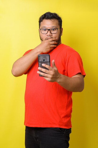 Fat asian guy with surprised expression covering mouth while looking to cell phone isolated on yello