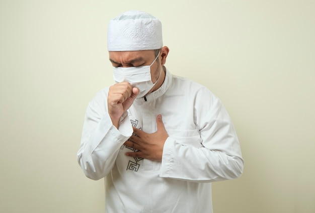 fat asian muslim men wearing a mask is coughing while closing his mouth using his hands